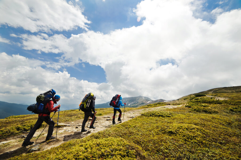 Skupina dospívajících mužů s batohy kráčející po horské cestě. Na obzoru jsou horské vrcholky, obloha je modrá a částečně zatažená. Mají před sebou ještě dlouhou cestu.
